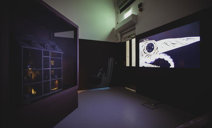 A dark and moody room with an eerie dolls house and animation playing on a tv screen.